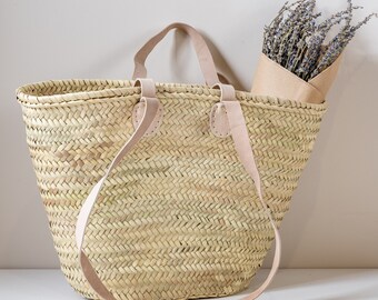 May include: A large, woven straw tote bag with leather straps. The bag is empty and has a light brown color. The bag is sitting on a white surface.