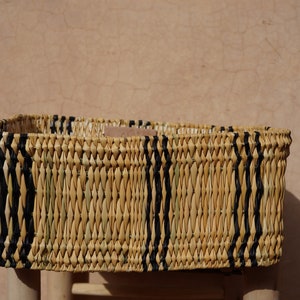 May include: Rectangular woven basket with natural and black vertical stripes. The basket is made of light-colored straw and sits on a light-colored wooden stool. The background is a textured, light brown wall.
