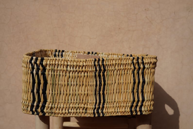 May include: Rectangular woven basket with natural and black vertical stripes. The basket is made of light-colored straw and sits on a light-colored wooden stool. The background is a textured, light brown wall.