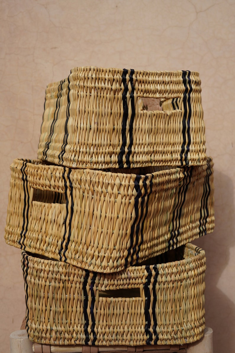 May include: Three rectangular woven storage baskets stacked on top of each other. The baskets are made of natural-colored straw with vertical black stripes and rectangular cut-out handles. The baskets are a neutral color and are suitable for home organization.