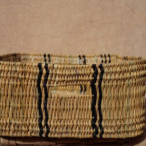 May include: Rectangular woven basket with a natural beige color and black accents. The basket has a rectangular shape with a handle cut out on each side. The basket is made of woven material.