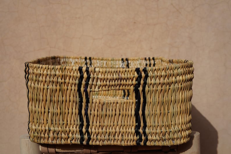 May include: Rectangular woven basket with a natural beige color and black accents. The basket has a rectangular shape with a handle cut out on each side. The basket is made of woven material.