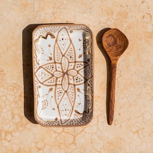Könnte beinhalten: Eine rechteckige Servierplatte aus weißem Keramik mit einem braunen und goldenen geometrischen Muster. Die Platte liegt neben einem Holzlöffel.