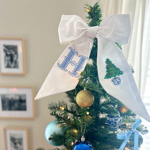 May include: A decorated Christmas tree with a large white bow as a tree topper. The bow has a blue letter "H" and a Christmas tree design. The tree is adorned with blue, gold, and light blue ornaments. The tree is decorated with lights.