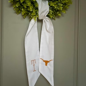 May include: A green wreath with a white ribbon featuring an embroidered orange letter "T" and a longhorn logo. The wreath is made of small, round green leaves.