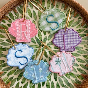 May include: Five embroidered seashell-shaped bag tags with gold-tone metal lobster clasps. Each tag features a unique fabric pattern and a large embroidered letter: a pink R, a green gingham S, a purple gingham L, a blue floral S, a blue floral I, and a pink palm tree J.
