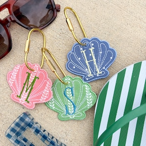 May include: Three seashell-shaped keychains in pink, green, and blue, each embroidered with a different letter. Gold-colored key rings are attached. Sunglasses and a green and white striped flip-flop are also visible.