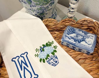 May include: A white fabric towel featuring an embroidered blue letter W and a detailed design of light blue hydrangea flowers in a blue and white patterned vase. The embroidery uses vibrant blue and green thread on a clean white background.