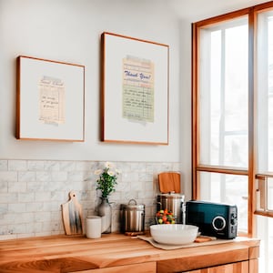 Puede incluir: Dos recibos vintage enmarcados con texto escrito a mano en tinta azul. El texto del recibo de la izquierda dice "Guest Check" y el texto del recibo de la derecha dice "Thank You!" Ambos recibos se muestran en una pared de azulejos blancos sobre una encimera de madera con electrodomésticos de cocina.