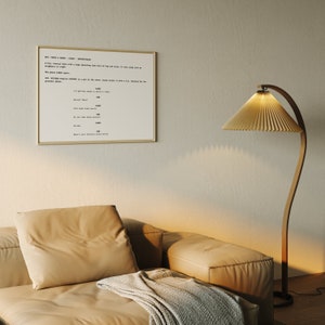 Könnte beinhalten: Ein beiges Ledersofa mit einer grauen Wolldecke. An der Wand hinter dem Sofa hängt ein gerahmtes Blatt Papier mit Text darauf. Eine Stehlampe aus Holz mit einem gefalteten, beigen Lampenschirm steht in der Ecke des Raumes.