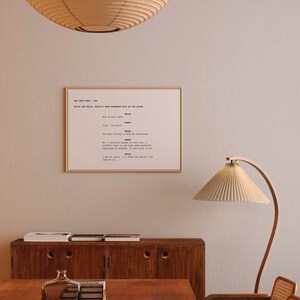 May include: A framed script print in a gold frame, hanging on a neutral wall. The script text reads "Bella and Harry's hand bandaged walk up the plank." A wooden cabinet and a lamp are in the foreground, creating a warm, inviting atmosphere.