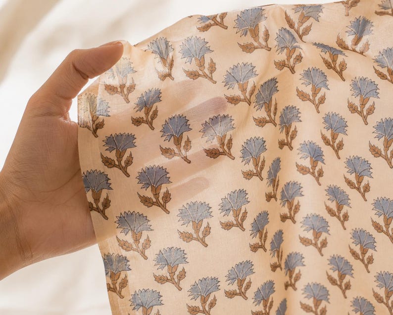 May include: A sheer, lightweight fabric featuring a repeating pattern of light blue flowers with brown stems and leaves on a pale beige background. The material is translucent, allowing light to pass through and revealing the texture of the weave.