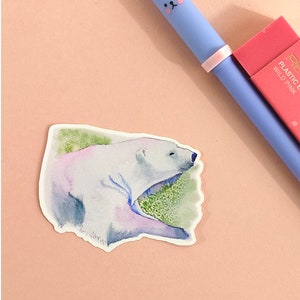 May include: A watercolor sticker of a polar bear lying down on a green background. The sticker is white with blue and purple watercolor accents.