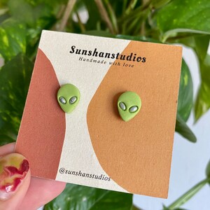 May include: A pair of green alien-shaped earrings with silver eyes. The earrings are displayed on a card with the text "Sunshanstudios Handmade with love" and "@sunshanstudios".