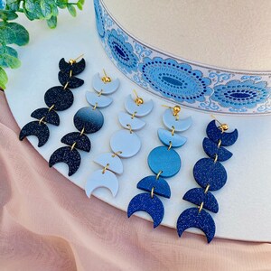 May include: Three pairs of dangle earrings featuring a crescent moon design. The earrings are made of polymer clay and come in black, white, and blue.