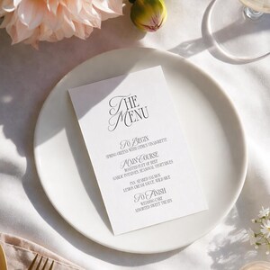 Op de afbeelding: Een trouwmenu op een wit bord met gouden bestek, kaarsen en bloemen. Het menu leest "THE MENU" en vermeldt gerechten als "Spring Greens" en "Roasted Filet of Beef."