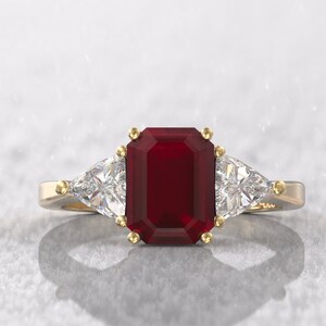 May include: A gold ring featuring a large, rectangular, deep red gemstone as the centerpiece, flanked by two smaller, triangular, clear gemstones. The ring has a classic design with a polished finish.