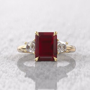 Puede incluir: Un anillo de oro con una gran gema rectangular de color rojo intenso como pieza central. Dos gemas claras, triangulares y más pequeñas flanquean la piedra principal. La banda del anillo es dorada y el engaste tiene garras doradas.
