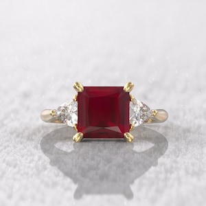 May include: A gold ring featuring a large, square-cut, deep red gemstone as the centerpiece, flanked by two smaller, triangular, clear gemstones. The ring's setting includes gold prongs and a gold band.