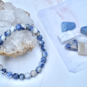 May include: A beaded bracelet with blue and white sodalite stones. The bracelet is next to a geode and several polished gemstones in shades of blue, white, and tan. The items are arranged on a white surface.
