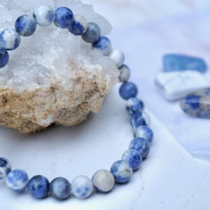 May include: A beaded bracelet made of blue and white sodalite stones. The bracelet is displayed next to a piece of rough quartz and several polished gemstones. The stones have a marbled appearance.