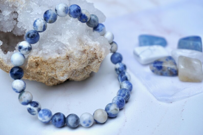 May include: A beaded bracelet made of blue and white sodalite stones. The bracelet is displayed next to a piece of rough quartz and several polished gemstones. The stones have a marbled appearance.