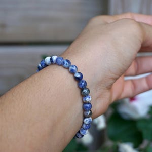May include: A beaded bracelet made of round, blue and white sodalite stones. The bracelet is worn on a wrist, showcasing the natural color variations of the stones. The bracelet is a piece of jewelry.