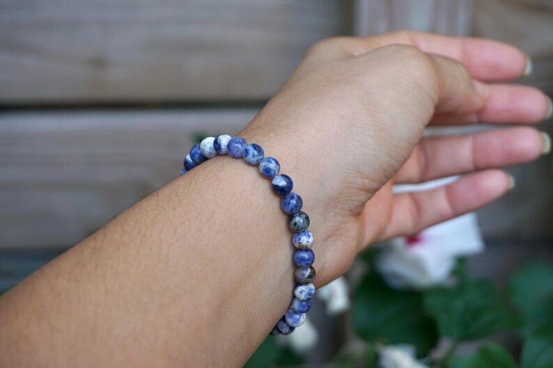 May include: A beaded bracelet made of round, blue and white sodalite stones. The bracelet is worn on a wrist, showcasing the natural color variations of the stones. The bracelet is a piece of jewelry.