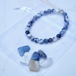 May include: A beaded bracelet with blue and white stones rests on a white drawstring bag. Several polished stones in shades of blue, white, and tan are scattered below the bracelet. The background is a light, neutral color.