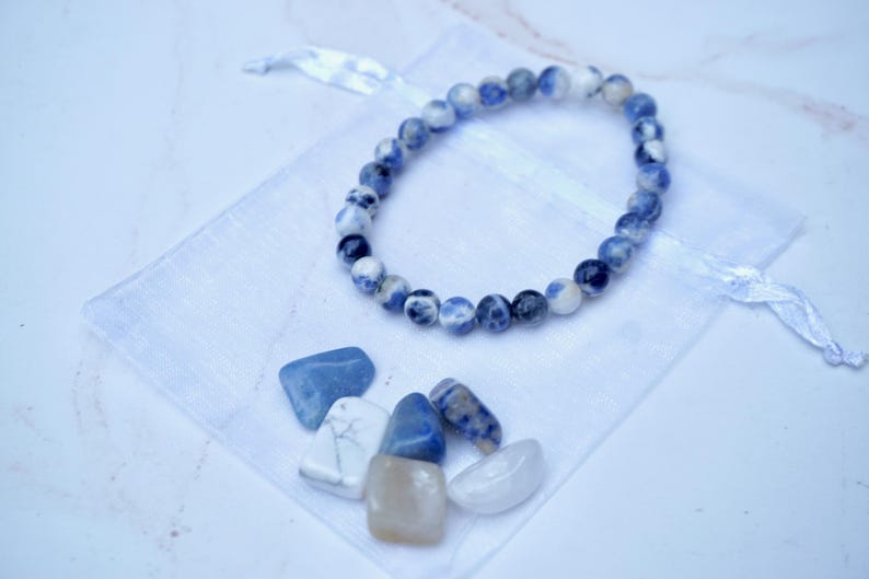 May include: A beaded bracelet with blue and white stones rests on a white drawstring bag. Several polished stones in shades of blue, white, and tan are scattered below the bracelet. The background is a light, neutral color.