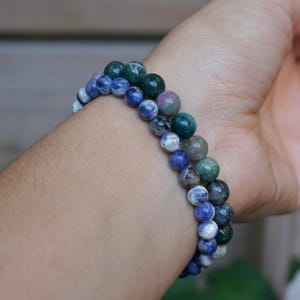 May include: Two beaded bracelets with blue, green, and white spherical beads are worn on a wrist. The bracelets are made of natural stone beads. The bracelets are a mix of colors and textures.