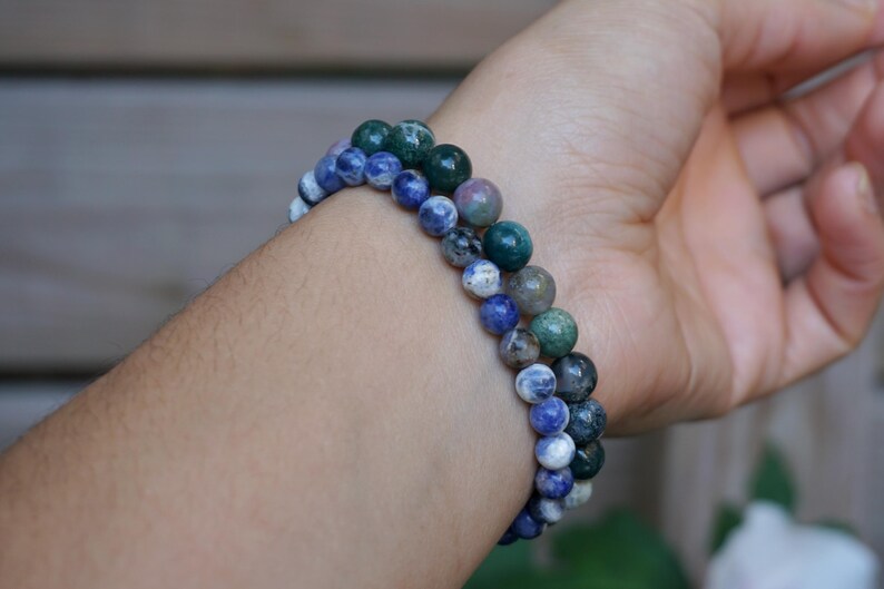 May include: Two beaded bracelets with blue, green, and white spherical beads are worn on a wrist. The bracelets are made of natural stone beads. The bracelets are a mix of colors and textures.