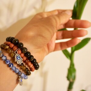 May include: Four beaded bracelets in shades of blue, brown, and black are stacked on a wrist. One bracelet has a silver charm with an eye design. The bracelets are made of various stones and beads.