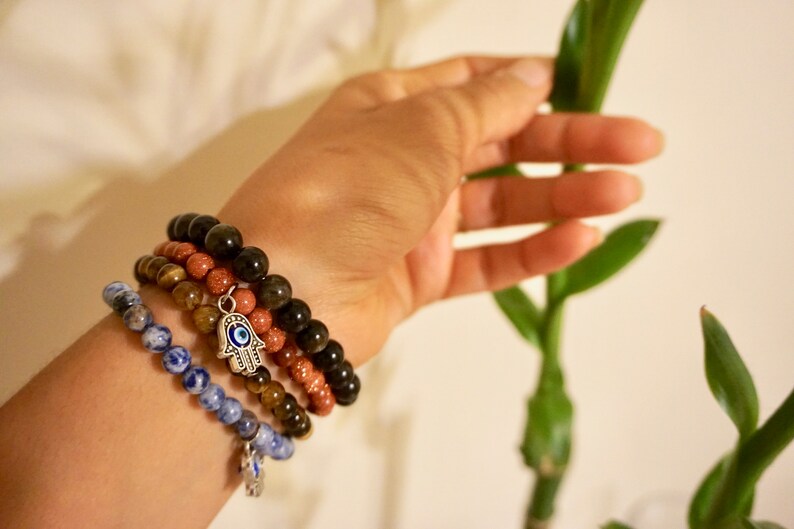 May include: Four beaded bracelets in shades of blue, brown, and black are stacked on a wrist. One bracelet has a silver charm with an eye design. The bracelets are made of various stones and beads.