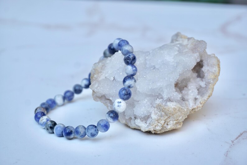 May include: A beaded bracelet with blue and white sodalite stones rests near a white crystal geode. The bracelet is strung with small, round beads, showcasing a mix of blue and white hues. The geode has a rough, beige exterior and a sparkling, crystalline interior.
