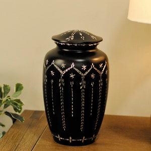 May include: Black cremation urn with a silver floral design. The urn has a lid and is sitting on a wooden surface.