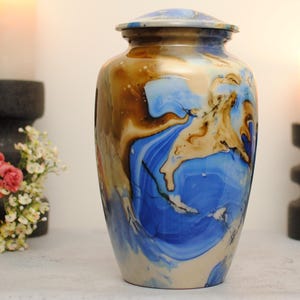 May include: A cylindrical metal cremation urn featuring a marbled finish with swirling patterns of deep blue, tan, and cream colours. The urn has a smooth, rounded lid that sits securely on top, creating a seamless silhouette for this vessel.