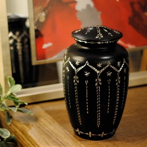 May include: Black urn with a silver design. The urn has a lid and is decorated with a geometric pattern.
