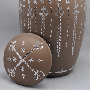 May include: A brown ceramic urn with a silver-colored design. The urn has a lid with a similar design. The urn is decorated with a repeating pattern of leaves and stars.