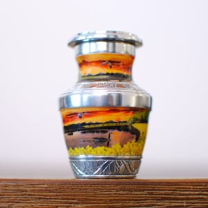 May include: A small silver urn with a painted landscape scene of a lake, trees, and a sunset. The urn has a silver lid and a decorative design around the base.