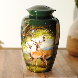 K&ouml;nnte beinhalten: Eine gr&uuml;ne Metallurne mit einem gemalten Bild von zwei Rehen in einer Waldlandschaft. Die Rehe sind braun mit wei&szlig;en Markierungen und stehen in einem grasbewachsenen Gebiet mit B&auml;umen und Blumen.