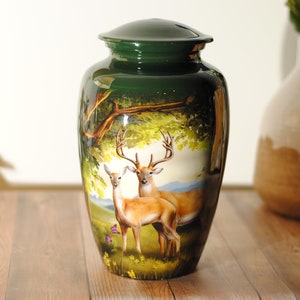 K&ouml;nnte beinhalten: Gr&uuml;ne Metallurne mit einer bemalten Szene von zwei Rehen in einer Waldlandschaft. Die Rehe sind braun mit wei&szlig;en Markierungen und stehen auf einer Lichtung mit gr&uuml;nen B&auml;umen und B&uuml;schen.