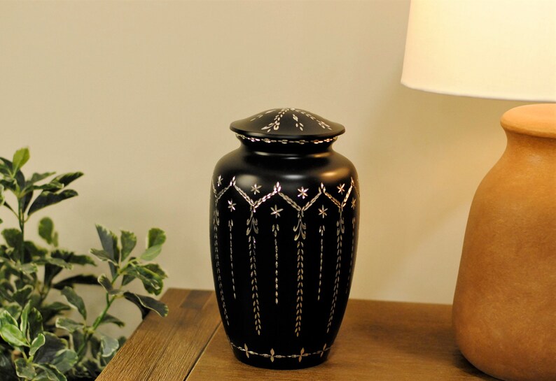 May include: Black cremation urn with a silver floral design. The urn has a lid and is sitting on a wooden surface.