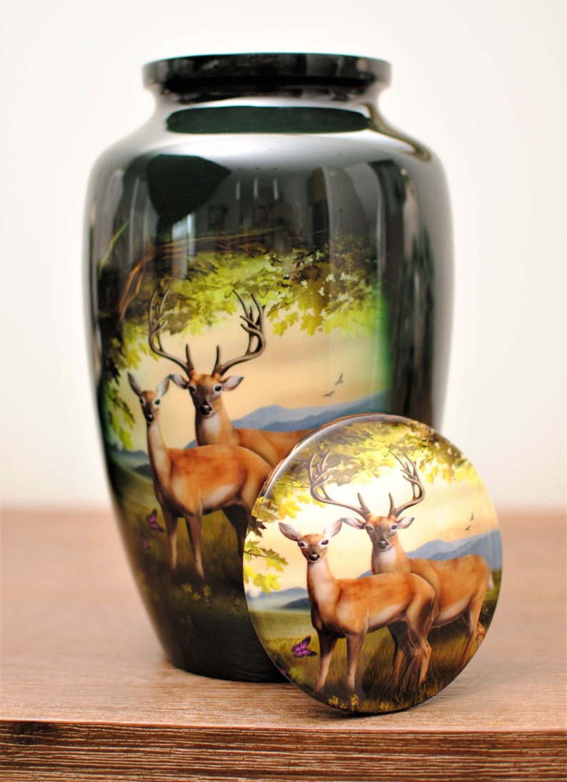 K&ouml;nnte beinhalten: Eine schwarze Keramikurne mit einer bemalten Szene von zwei Hirschen in einer Waldlandschaft. Der Deckel der Urne zeigt die gleiche Szene.