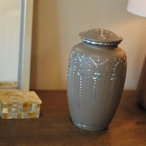 May include: A gray ceramic urn with a silver design. The urn has a lid and is decorated with a silver floral pattern.