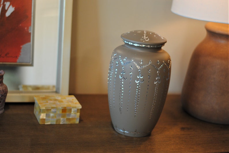 May include: A gray ceramic urn with a silver design. The urn has a lid and is decorated with a silver floral pattern.