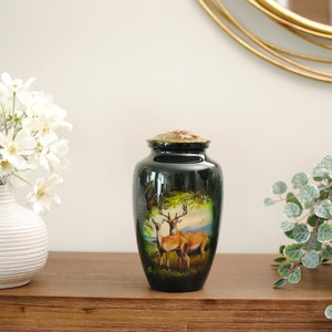 K&ouml;nnte beinhalten: Eine schwarze Metallurne mit einem goldenen Deckel und einem gemalten Bild von zwei Rehen in einer Waldlandschaft. Die Urne steht auf einer Holzoberfl&auml;che mit wei&szlig;en Blumen in einer Vase und einer gr&uuml;nen Pflanze in einem Topf.