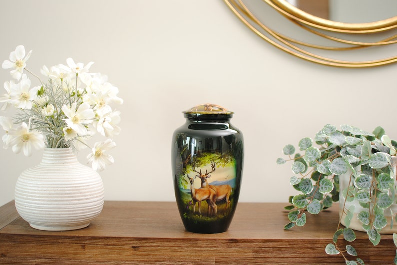 K&ouml;nnte beinhalten: Eine schwarze Metallurne mit einem goldenen Deckel und einem gemalten Bild von zwei Rehen in einer Waldlandschaft. Die Urne steht auf einer Holzoberfl&auml;che mit wei&szlig;en Blumen in einer Vase und einer gr&uuml;nen Pflanze in einem Topf.