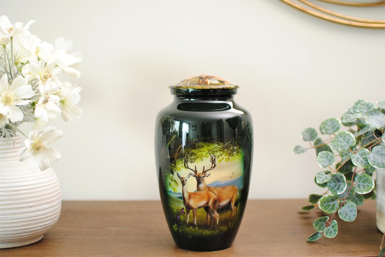 K&ouml;nnte beinhalten: Eine schwarze Metallurne mit einem goldenen Deckel und einer bemalten Szene mit zwei Hirschen in einem Wald. Die Urne steht auf einer Holzoberfl&auml;che.