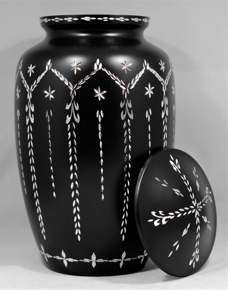 May include: Black cremation urn with a silver floral design. The urn has a lid with a matching design.